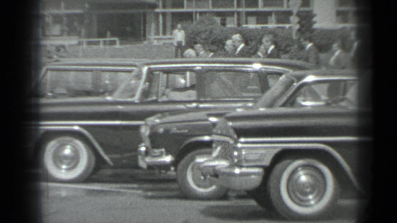 1983 Moscow home-movie frame showing a row of dark cars and suited men near a building, seen through a black-and-white 8mm gate.