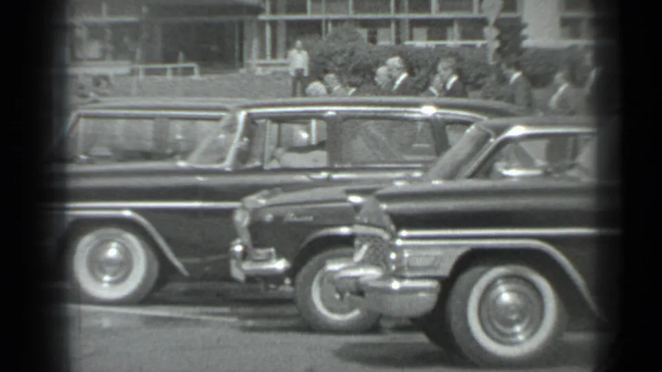 1983 Moscow home-movie frame showing a row of dark cars and suited men near a building, seen through a black-and-white 8mm gate.