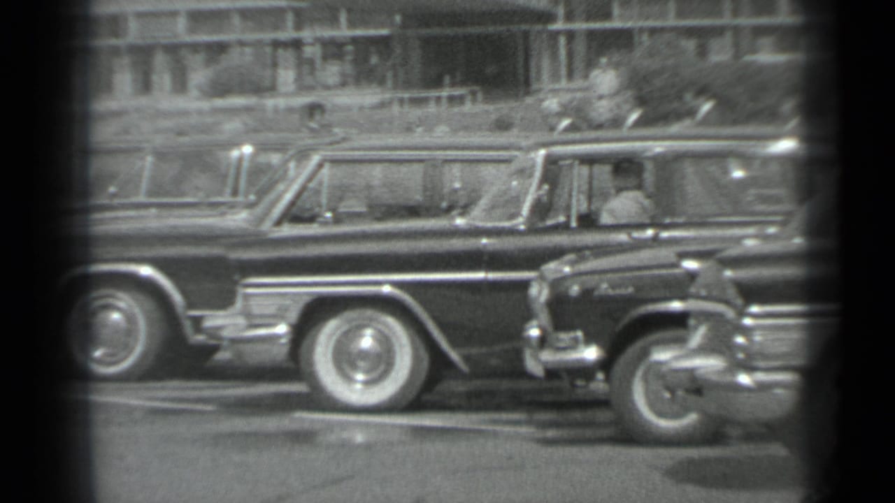Black-and-white 8mm frame from Moscow in 1983 showing parked official-looking cars and suited men along a city street.
