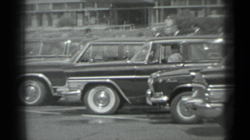 Black-and-white 8mm frame from Moscow in 1983 showing parked official-looking cars and suited men along a city street.