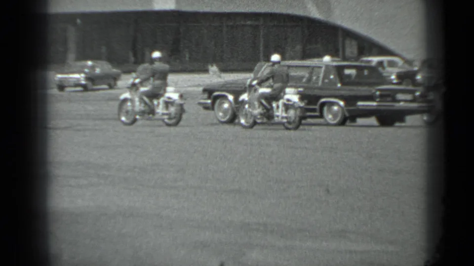 A dark sedan or limousine moves with motorcycle police nearby in a Soviet-era Moscow street scene, per supplied metadata.
