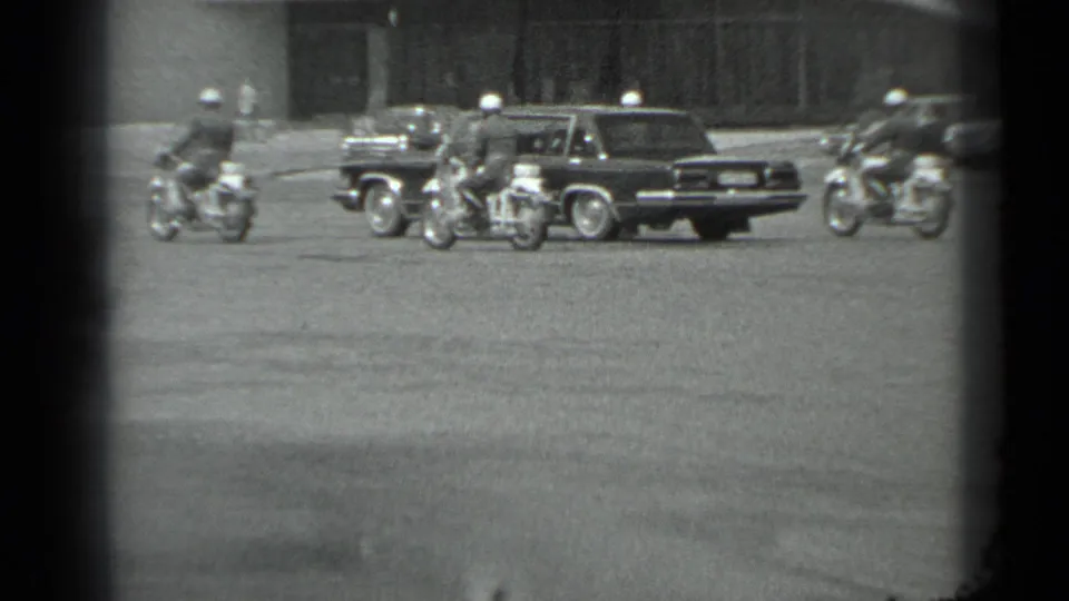 Motorcycle officers ride around a dark official-looking car on a broad Moscow roadway, with buildings behind them.