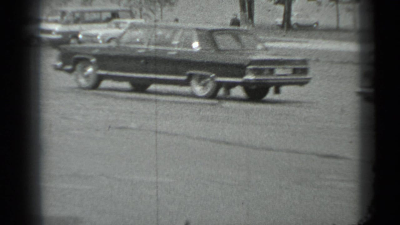 Black-and-white 8mm frame of a large sedan on a Moscow street in the 1983 home-movie sequence.