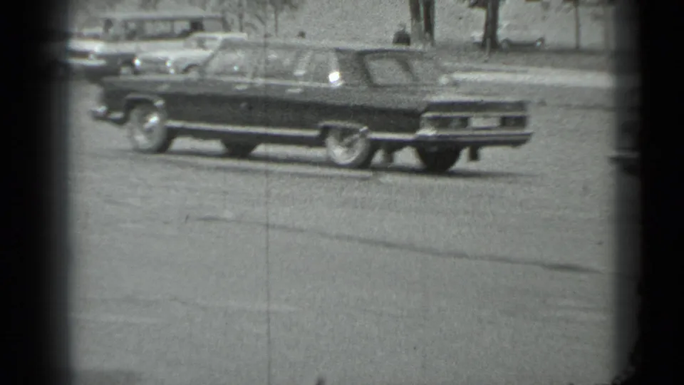 Black-and-white 8mm frame of a large sedan on a Moscow street in the 1983 home-movie sequence.