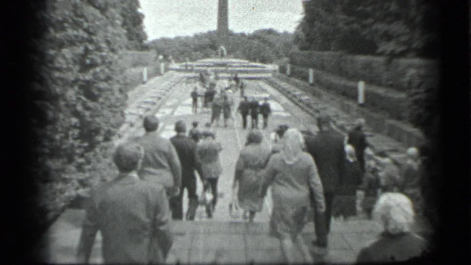 Civilians walk up a broad landscaped path toward a tall distant monument in the 1983 Moscow home movie.