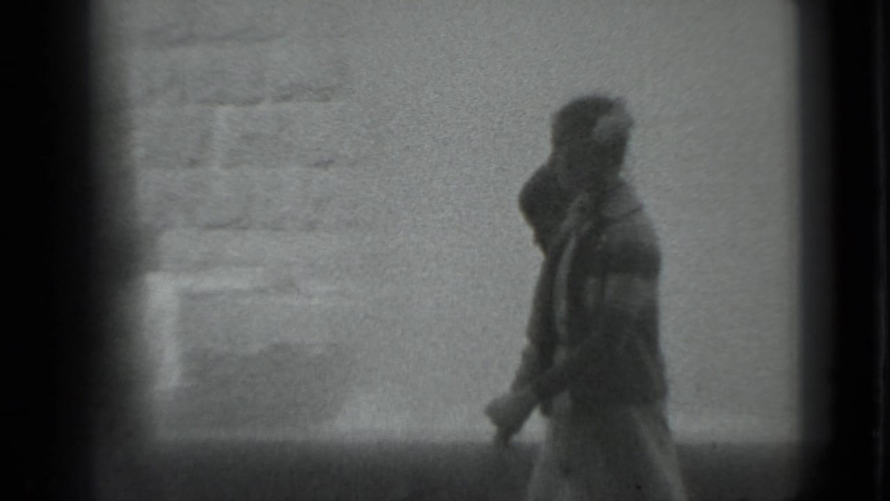 A blurred figure walks past a pale textured wall in a grainy black-and-white frame.