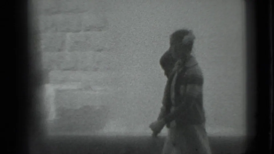 A solitary pedestrian appears to walk past a textured wall in 1983 Moscow, softened by motion blur and 8mm grain.