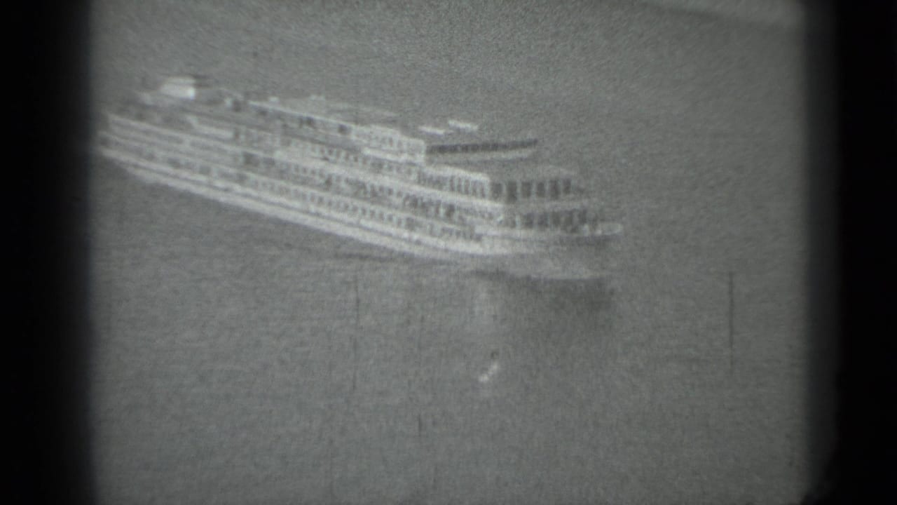 Distant multi-deck passenger ship moving across calm water within dark film borders.