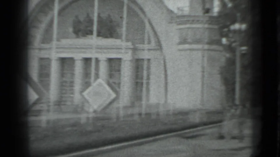 1983 Moscow 8mm frame showing the arched facade from a slight side angle, with columns, flagpoles, a sign, and a walkway along the right edge.