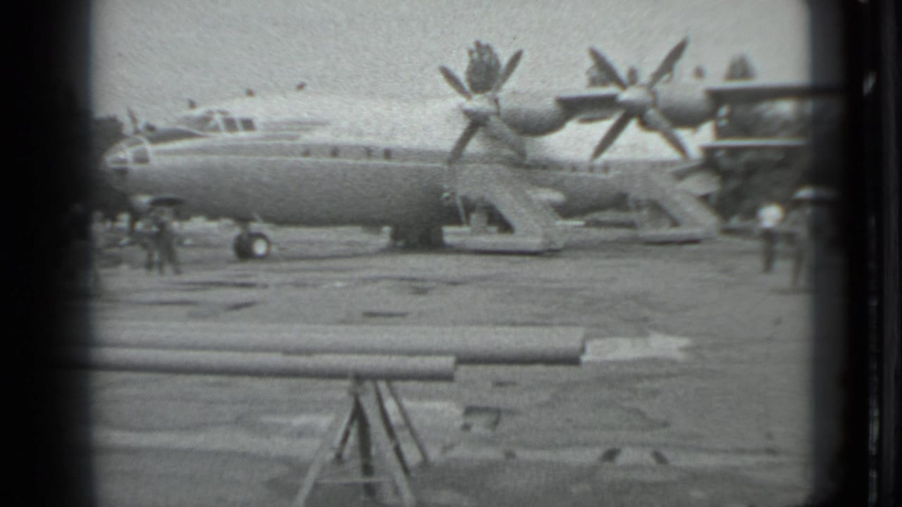 Propeller aircraft seen from the side with people on the tarmac and dark film edges.