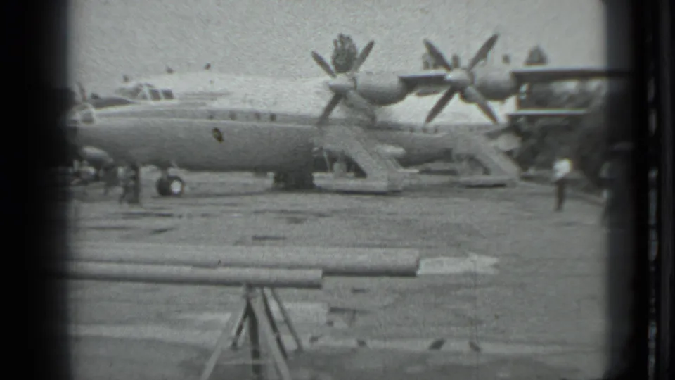 Grainy 8mm frame of the Moscow tarmac showing the large propeller aircraft, nearby people, and a person moving at the right edge.