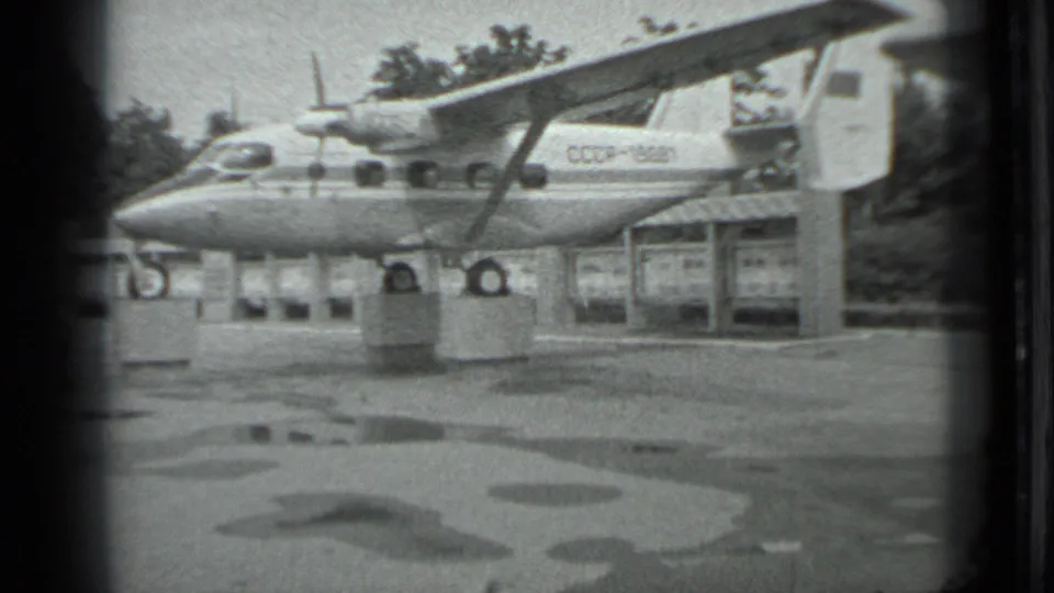 The 8mm frame holds on the Soviet-marked aircraft, concrete display supports, puddles, and background shelter.