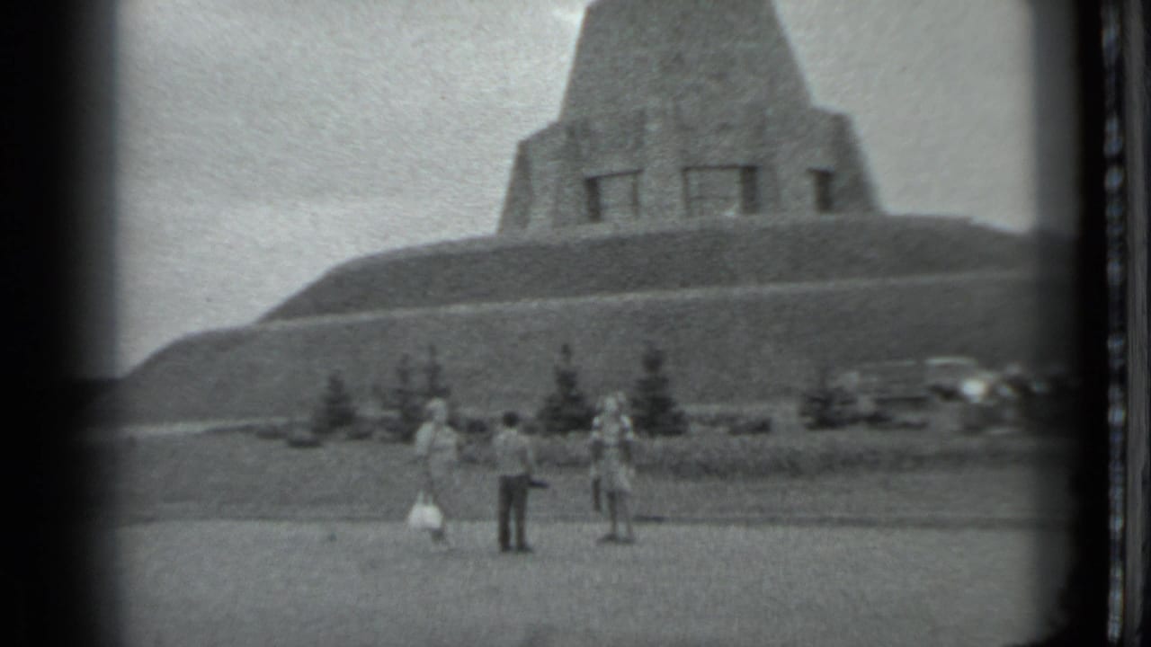 Three people stand near a large concrete monument on a grassy rise.