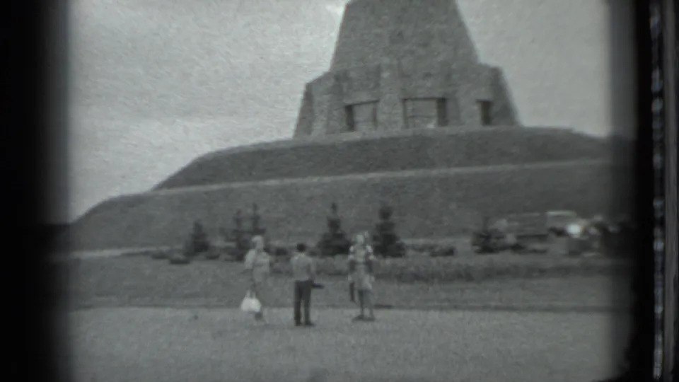 In 1983 Moscow home-movie footage, three adults stand before a large concrete monument with trees and parked cars nearby.