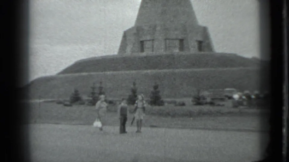 The group remains near the monument base in grainy 8mm footage, with the concrete mass rising above a grassy mound.