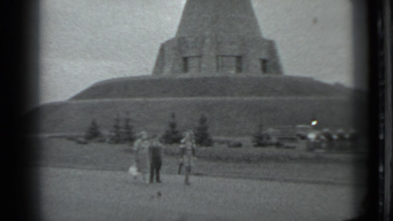 1983 Moscow home-movie frame showing people on a grassy area with a large pyramid-shaped hillside monument behind them.