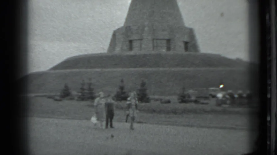 1983 Moscow home-movie frame showing people on a grassy area with a large pyramid-shaped hillside monument behind them.