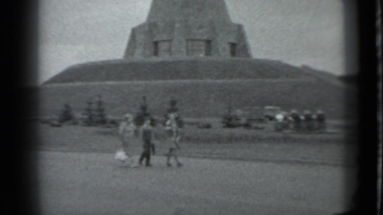 Three people move across a grassy foreground with a monument and cars behind them.