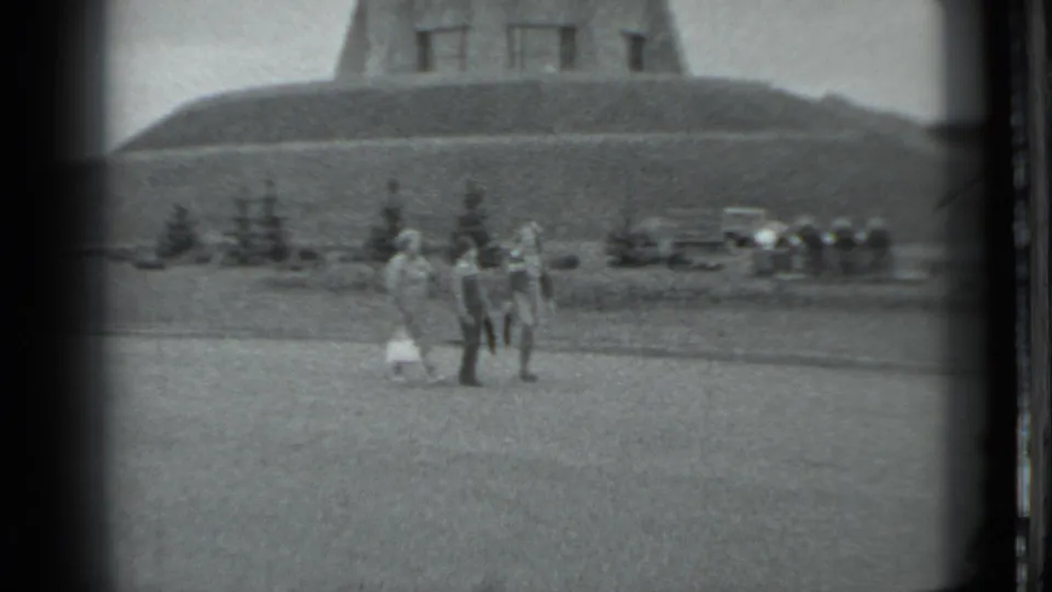 1983 Moscow home-movie still showing companions gathered on grass, with the hillside monument partly cropped above them.