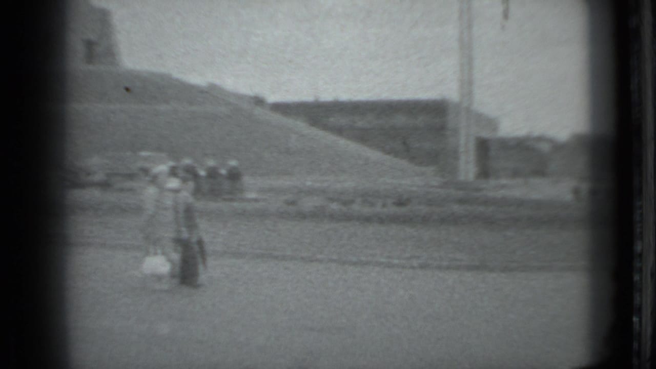 In 1983 Moscow footage, distant pedestrians walk across a broad paved plaza near monumental concrete forms and tall vertical poles.