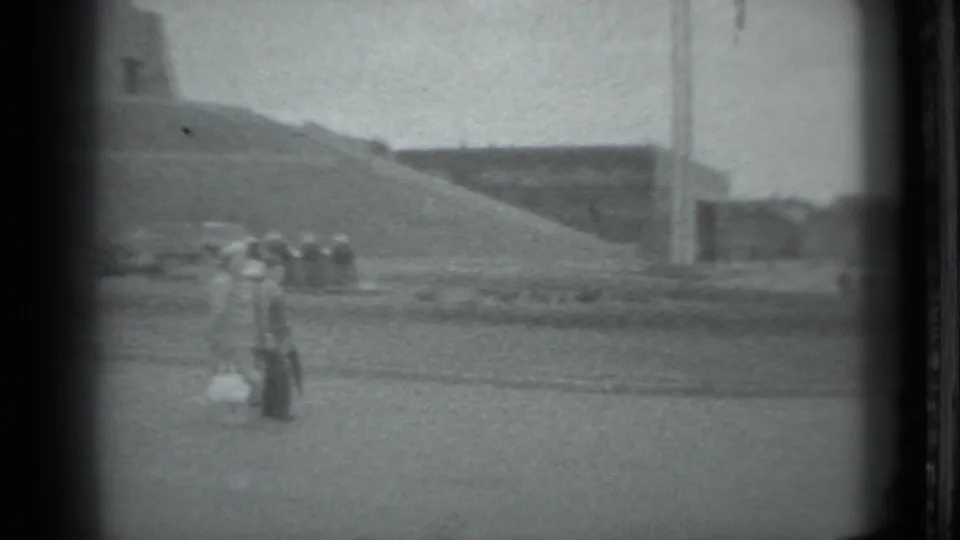 In 1983 Moscow footage, distant pedestrians walk across a broad paved plaza near monumental concrete forms and tall vertical poles.
