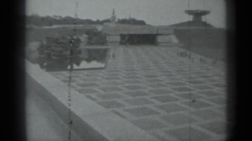 A patterned Moscow plaza is seen with low walls, distant pedestrians, vertical poles, and large modernist concrete structures.