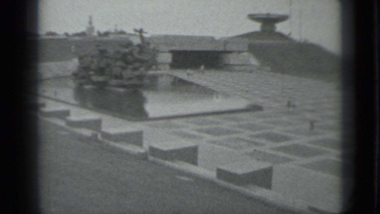 Wide black-and-white view of a memorial plaza with a central sculptural fountain and mound beyond.
