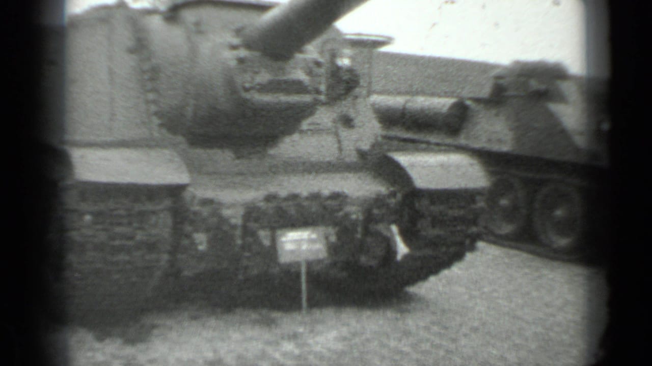 1983 Moscow home-movie frame showing a Soviet-era tank or armored vehicle on exhibition, with gun barrel, tracks, and placard visible.