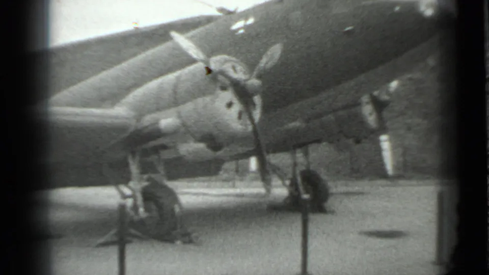 1983 Moscow home-movie frame showing a propeller military aircraft on outdoor display, with engine, wing, landing gear, and barriers visible.