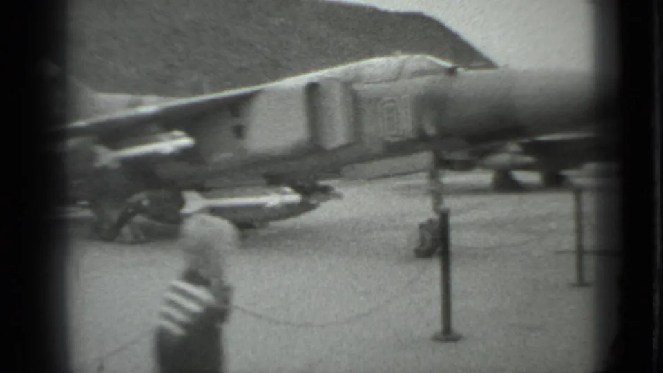 1983 Moscow 8mm frame showing a parked military jet fighter from the side, with barriers and a foreground post partly obscuring the view.