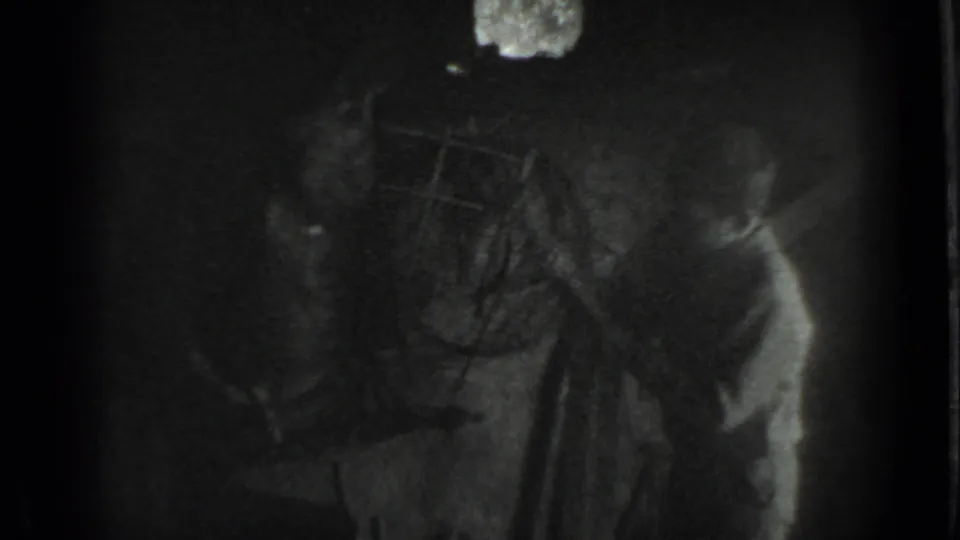 1983 Moscow home-movie frame showing a boy near an anvil or work surface in a dim workshop with heavy grain and deep shadows.