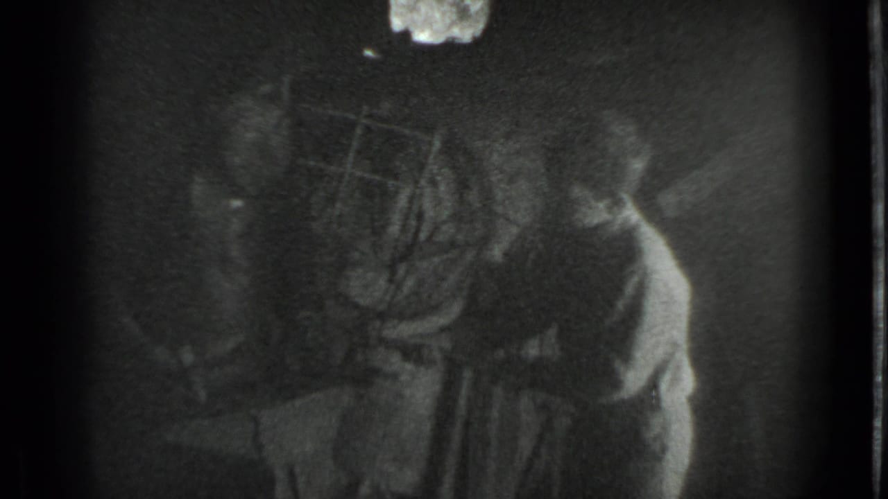 Grainy 8mm frame from the Moscow workshop scene, with the boy leaning near the anvil and tools partly obscured by shadow.