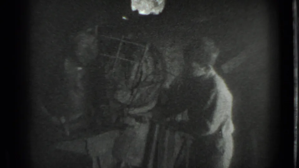 Grainy 8mm frame from the Moscow workshop scene, with the boy leaning near the anvil and tools partly obscured by shadow.