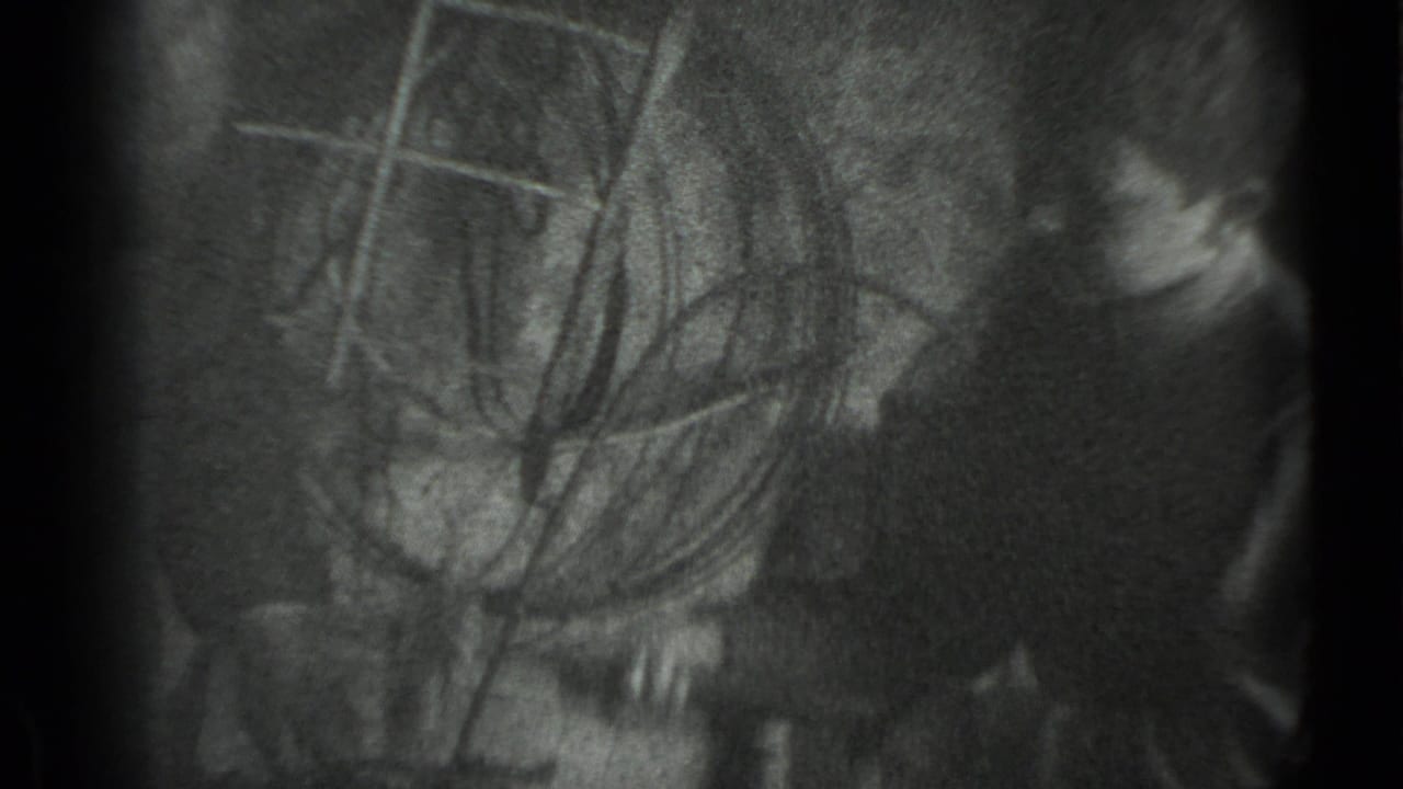Grainy monochrome frame with a partially visible person and tool-like lines in a dark interior.