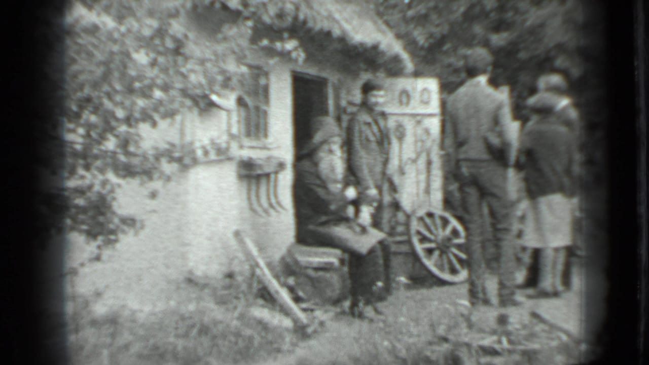 1983 Moscow-region home-movie frame showing adults outside a rustic cottage with a seated bearded man, horseshoes, and a wagon wheel.