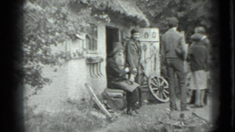 1983 Moscow-region home-movie frame showing adults outside a rustic cottage with a seated bearded man, horseshoes, and a wagon wheel.