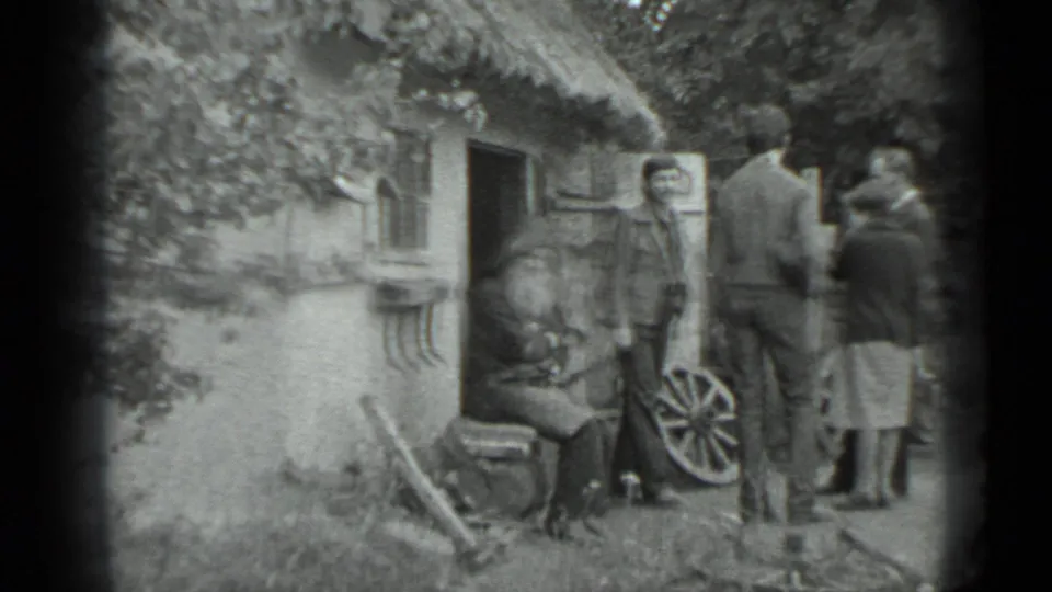 Adults stand beside a rustic cottage doorway in the Moscow 1983 footage, near a seated man and a wagon wheel display.