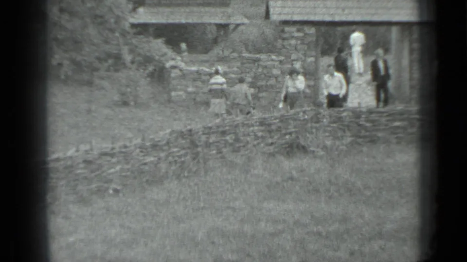 In the supplied 1983 Moscow, Russia home-movie frame, several people move near stone walls and older village buildings in soft 8mm focus.