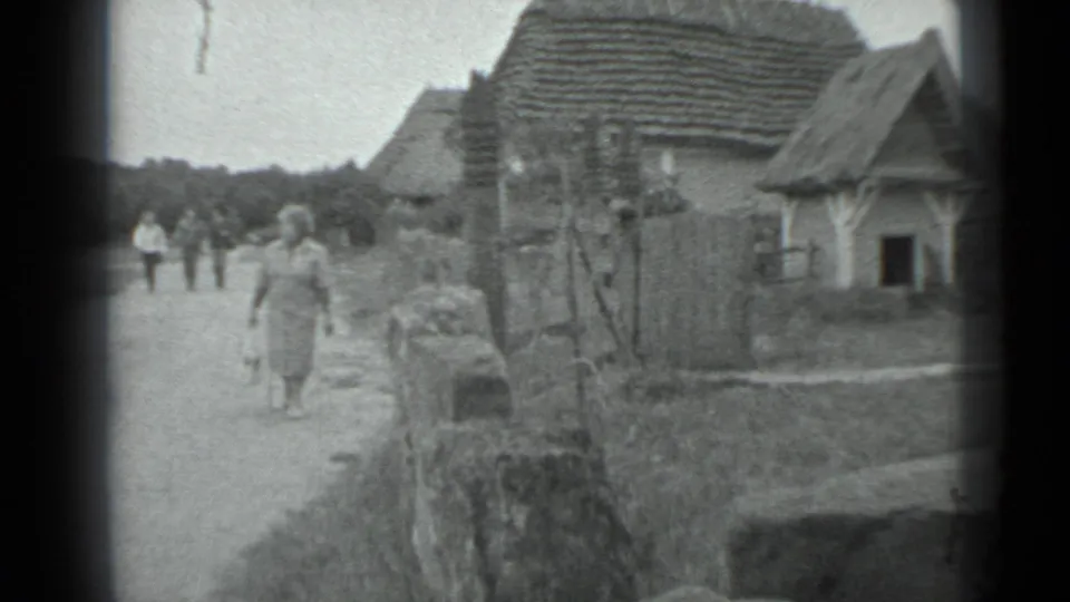 The frame shows a woman on a village road, older houses to the right, and distant figures in a soft black-and-white 8mm scan.