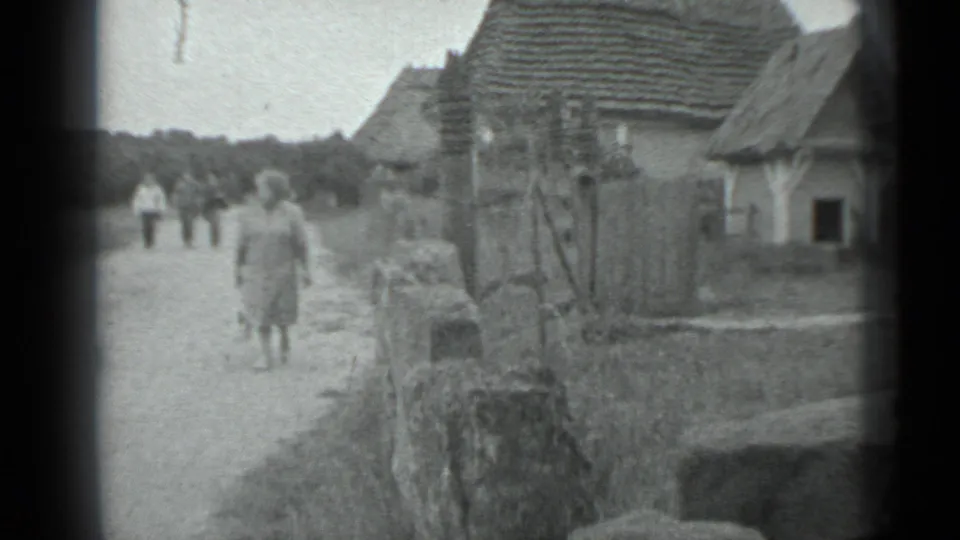 In 1983 Moscow-area rural footage, a woman in a light dress walks toward the camera along a dirt road near village houses.