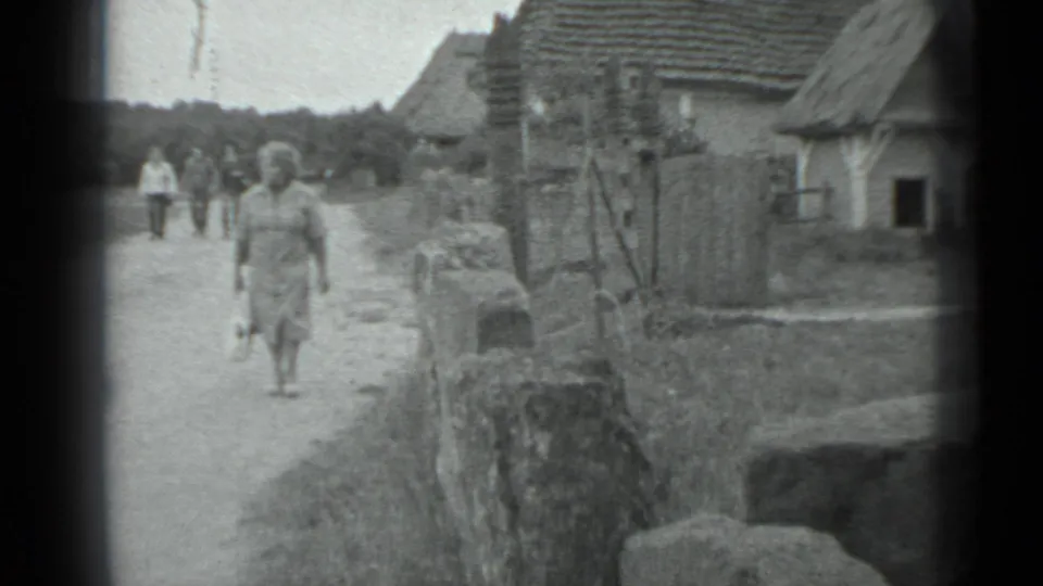 A woman walks forward on a rural dirt path near Moscow in 1983, passing a low wall and village houses as people follow behind.