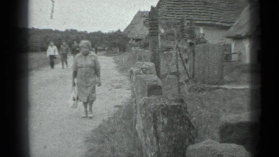 The 8mm frame shows the woman nearer to camera on a village road, with rustic houses, a wall, and distant pedestrians visible.