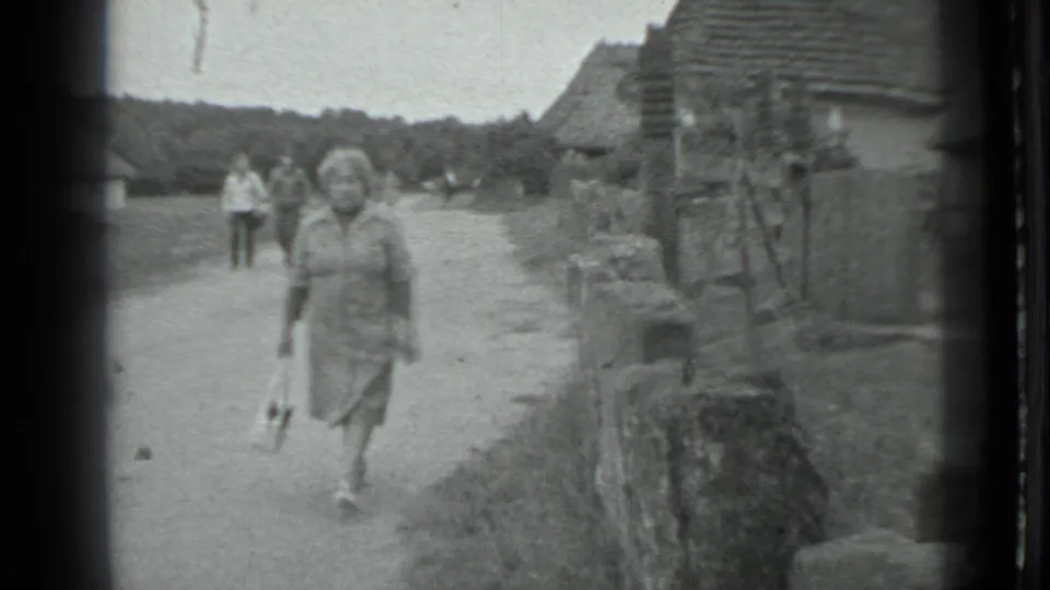 Near the end of the 1983 rural Moscow clip, the woman passes closer to camera while houses, a wall, and walkers remain behind her.