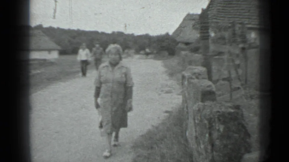 In a 1983 Moscow-area rural village scene, a curly-haired woman in a dress walks toward the camera on a dirt road with huts and people behind her.