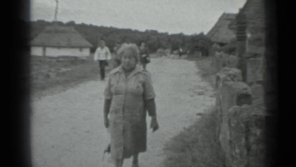The woman continues walking toward the camera in the rural Moscow-area setting, with low walls, huts, open ground, and background pedestrians visible.