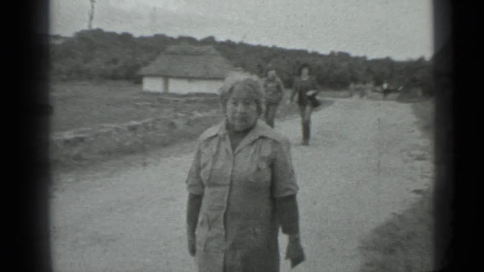 A closer 8mm frame shows the woman nearing the camera on the rural road, with open grass, a low white building, trees, and pedestrians in the background.
