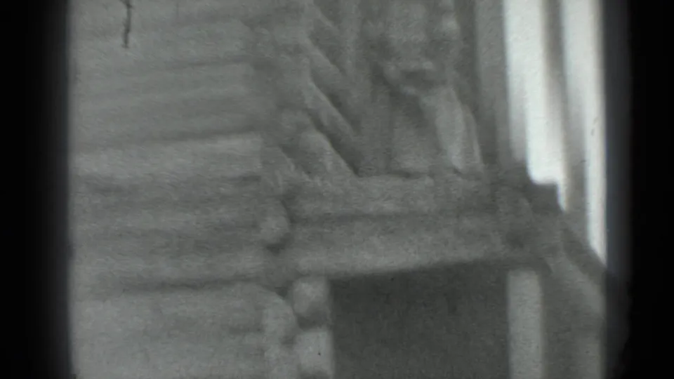 Grainy 1983 Moscow 8mm frame showing a log cabin wall and steep wooden ladder before the man is clearly visible.