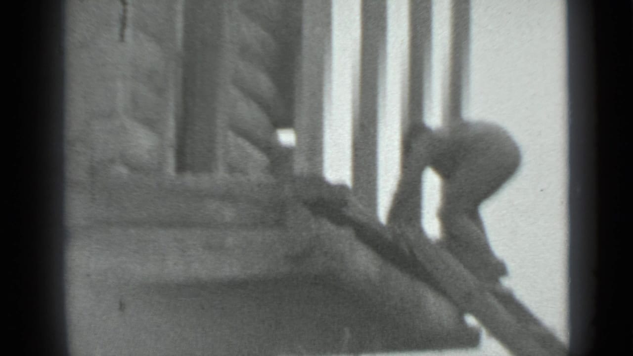 A man in dark clothing leans over a steep wooden ladder beside a log cabin in grainy 1983 Moscow home-movie footage.