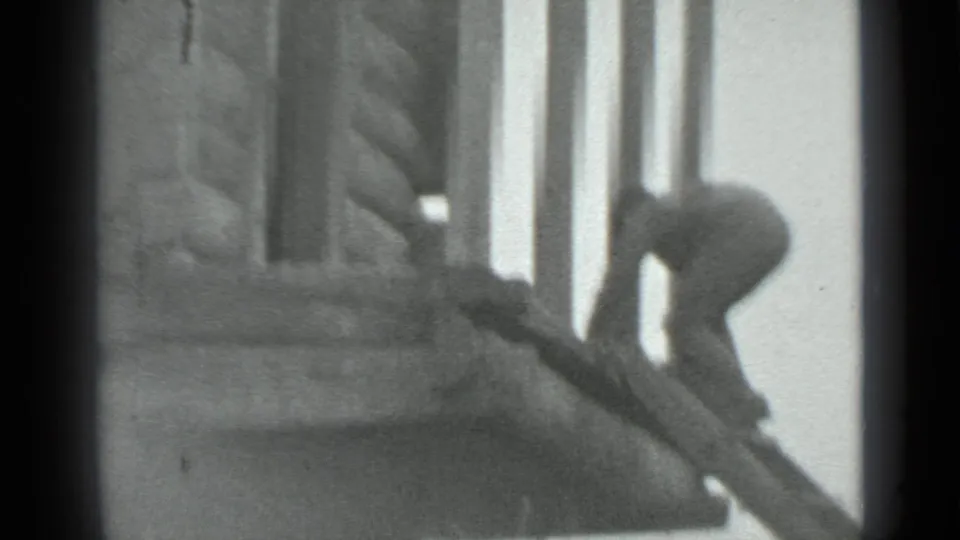 A man in dark clothing leans over a steep wooden ladder beside a log cabin in grainy 1983 Moscow home-movie footage.