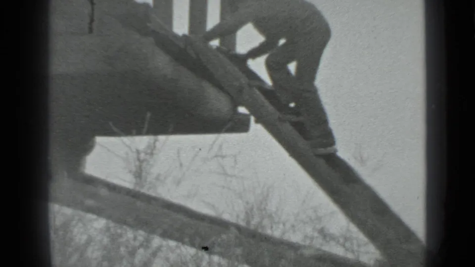 The man crouches and shifts his weight on the steep ladder against the log cabin, filmed from below in black-and-white 8mm.